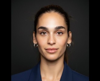 Professional head-and-shoulders portrait of a woman with dark hair pulled back, wearing a blue blazer, looking at the camera against a dark background.