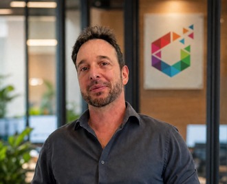 Man with short dark hair and stubble, wearing a dark shirt, standing in a modern office with a colorful geometric logo behind him.