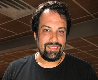 Man with dark hair and beard wearing a black t-shirt, smiling at the camera in an indoor setting.
